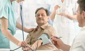 Starszy pan z wąsami ma mierzone **dbp ciśnienie**. Pielęgniarka delikatnie kładzie dłoń na jego ramieniu.