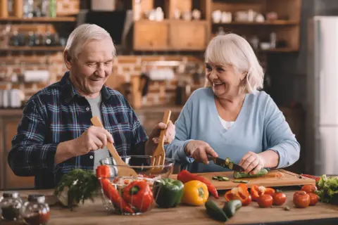 Uśmiechnięci seniorzy przygotowują zdrową sałatkę. To przykład, jak dbać o dietę seniora, ciesząc się wspólnym gotowaniem.
